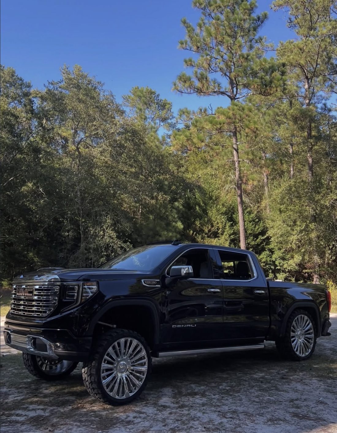 Black GMC Denali crew cab with a tall stance and chrome wheels, parked in front of pine trees