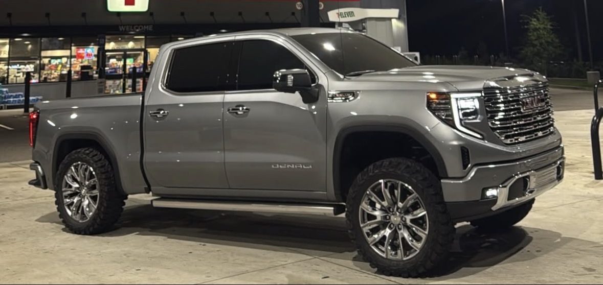 Grey GMC Denali pickup with polished wheels and mud-terrain tires, parked at night under bright canopy lights